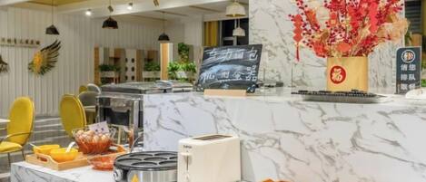 Un espace d’accueil d’hôtel avec un comptoir en marbre, un panier de fruits, un grille-pain et un vase rempli de fleurs rouges.