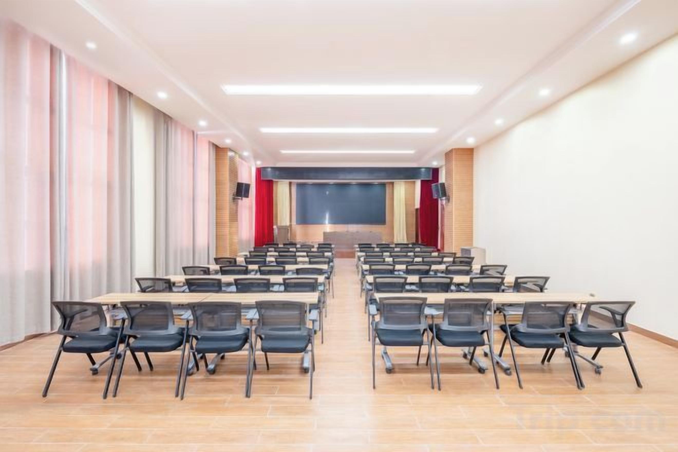 Un salón de conferencias con filas de mesas y sillas, un escenario con una pantalla y un ventanal grande en un lateral.
