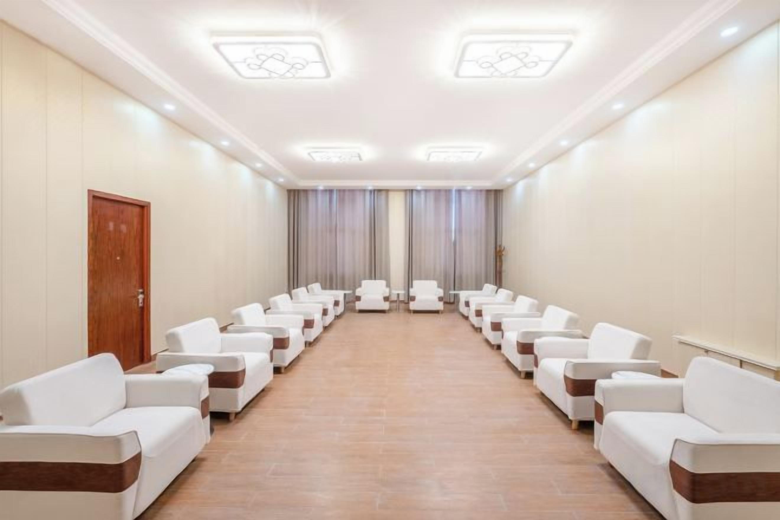 A hotel room with a long row of white sofas facing a window with curtains, a wooden door on the left, and a tiled floor.