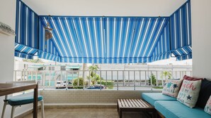 A balcony with a blue-striped awning, a wooden bench, a cushioned sofa, and a view of the street. - Oceanview Apartment by Playa Azul - walk to shore, snorkel, swim, sun, unwind. (Luquillo)