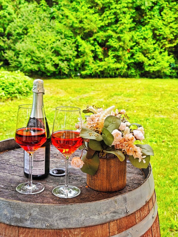 Un barril de madera con una botella de vino, dos copas de vino y un pequeño florero con flores sobre un paisaje natural cubierto de hierba.