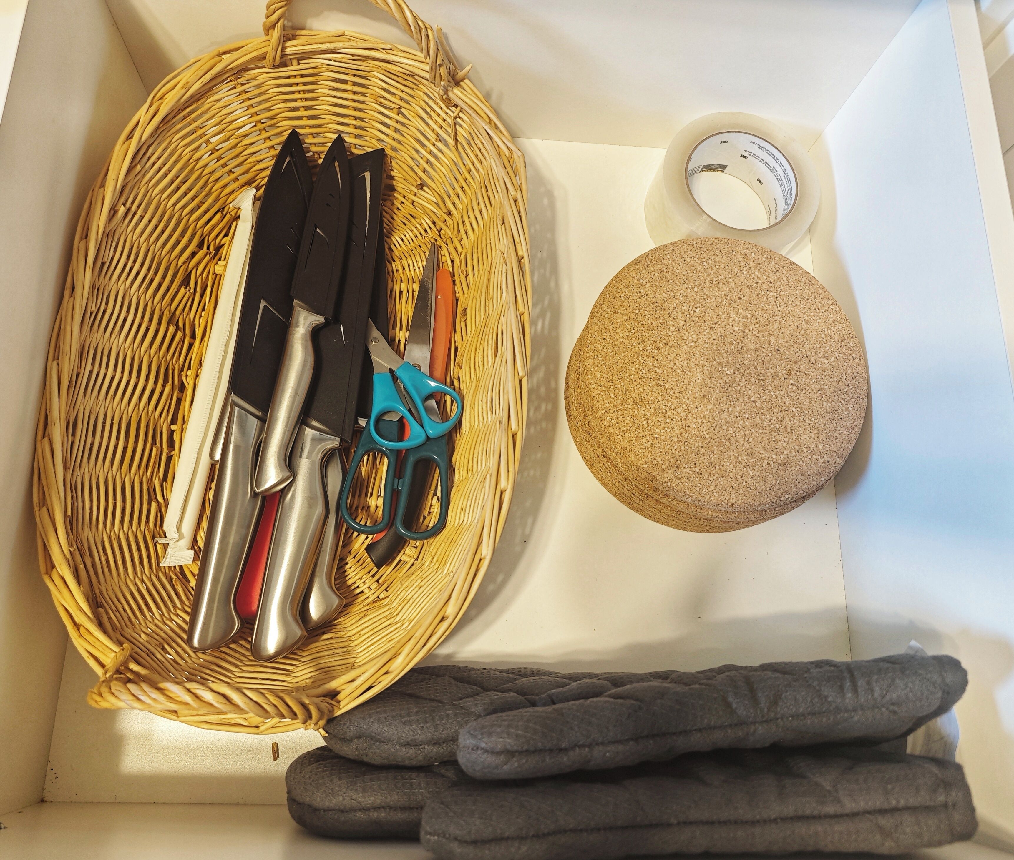 A kitchen drawer with a wicker basket containing kitchen utensils, a cork, and a roll of tape.