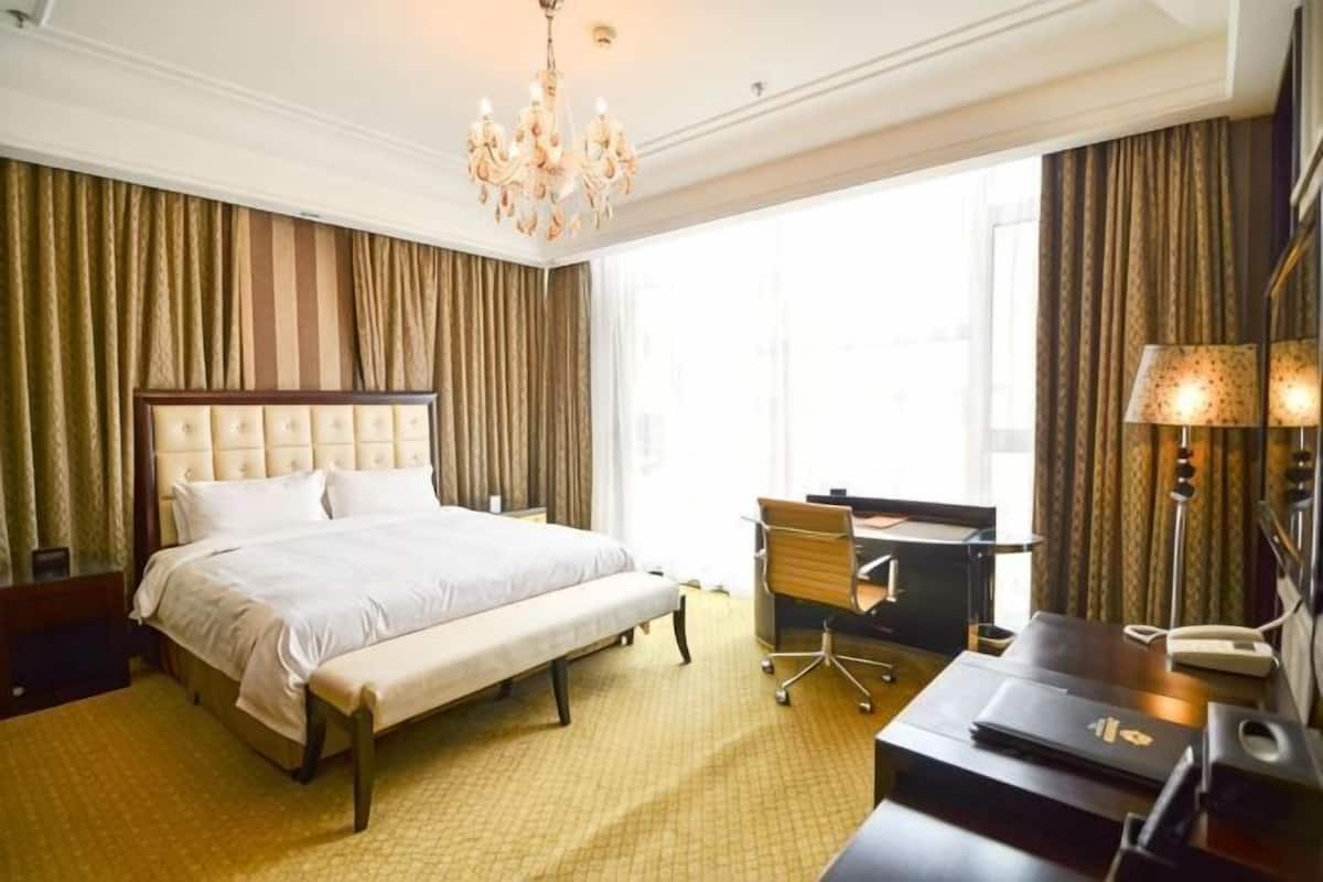 Ein Hotelzimmer mit einem großen Bett, einem Schreibtisch mit Stuhl, einer Schreibtischlampe und einem Fenster mit Vorhängen.