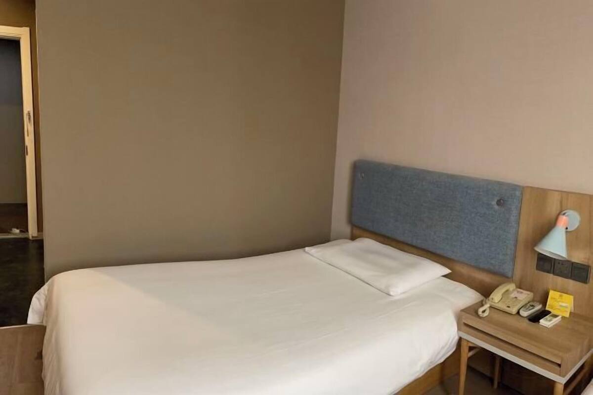 Una cama individual con cabecera de madera, una mesita de noche con lámpara, un teléfono y una tarjeta pequeña.