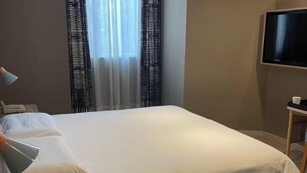 A neatly made bed with white linens, a bedside lamp, a television on the wall, and a window with patterned curtains. - For example, hotels (Shanghai)