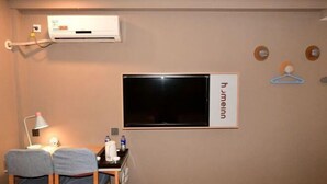 A room with a flat-screen TV mounted on the wall, a desk with a lamp, two chairs, and a small table with a water bottle.