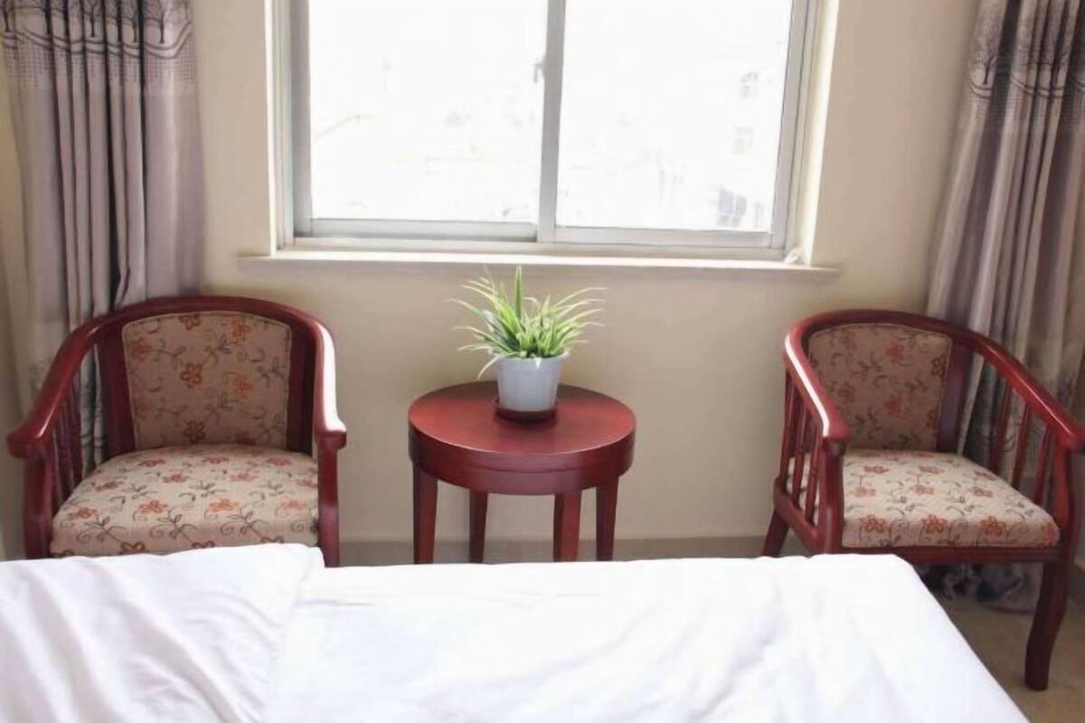 A room with a bed, two floral-patterned chairs, a small table with a plant, and a window with curtains.