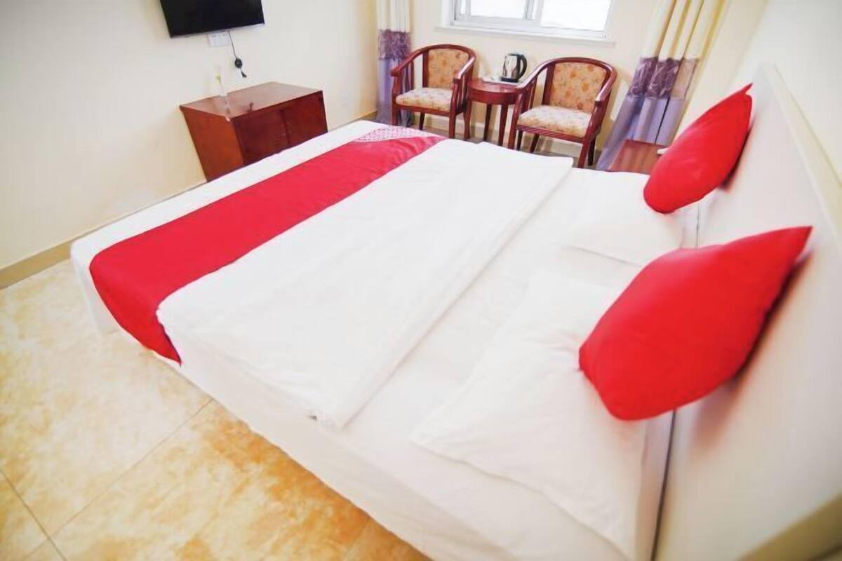 A hotel room with a bed, a small wooden side table, two chairs, and a TV mounted on the wall.