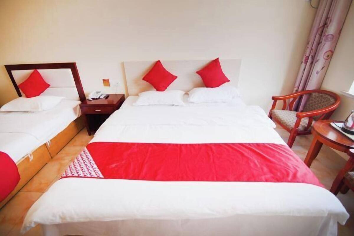 A hotel room with two beds, each with red and white bedding. There is a wooden nightstand with a phone and a lamp between the beds. A wooden chair and a small table are visible in the corner.