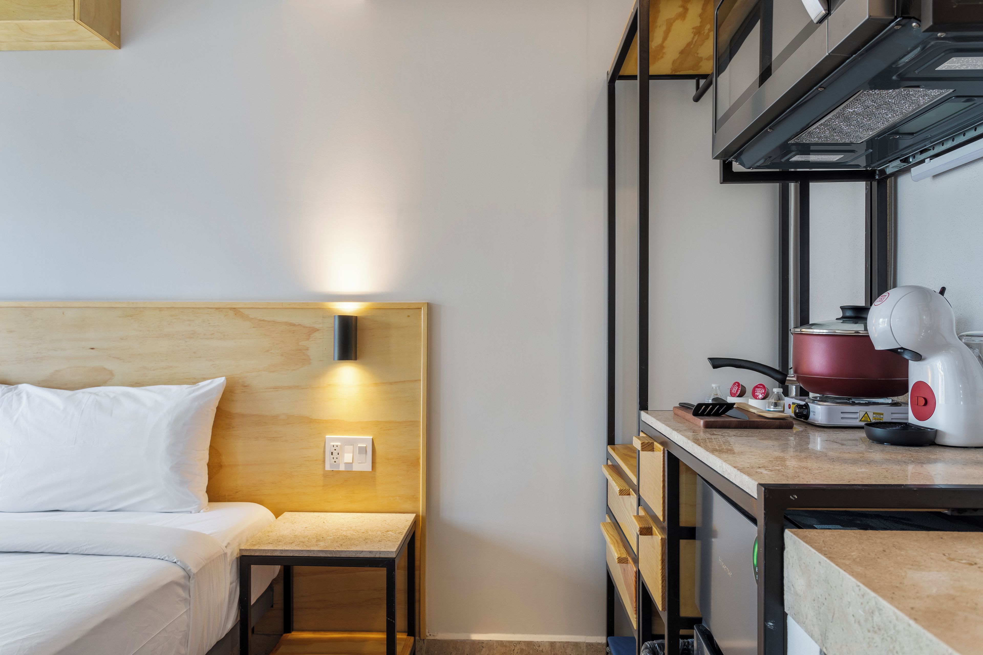 Habitació d'hotel compacta amb llit, taula de nit petita, cuineta equipada amb cafetera i fogó.