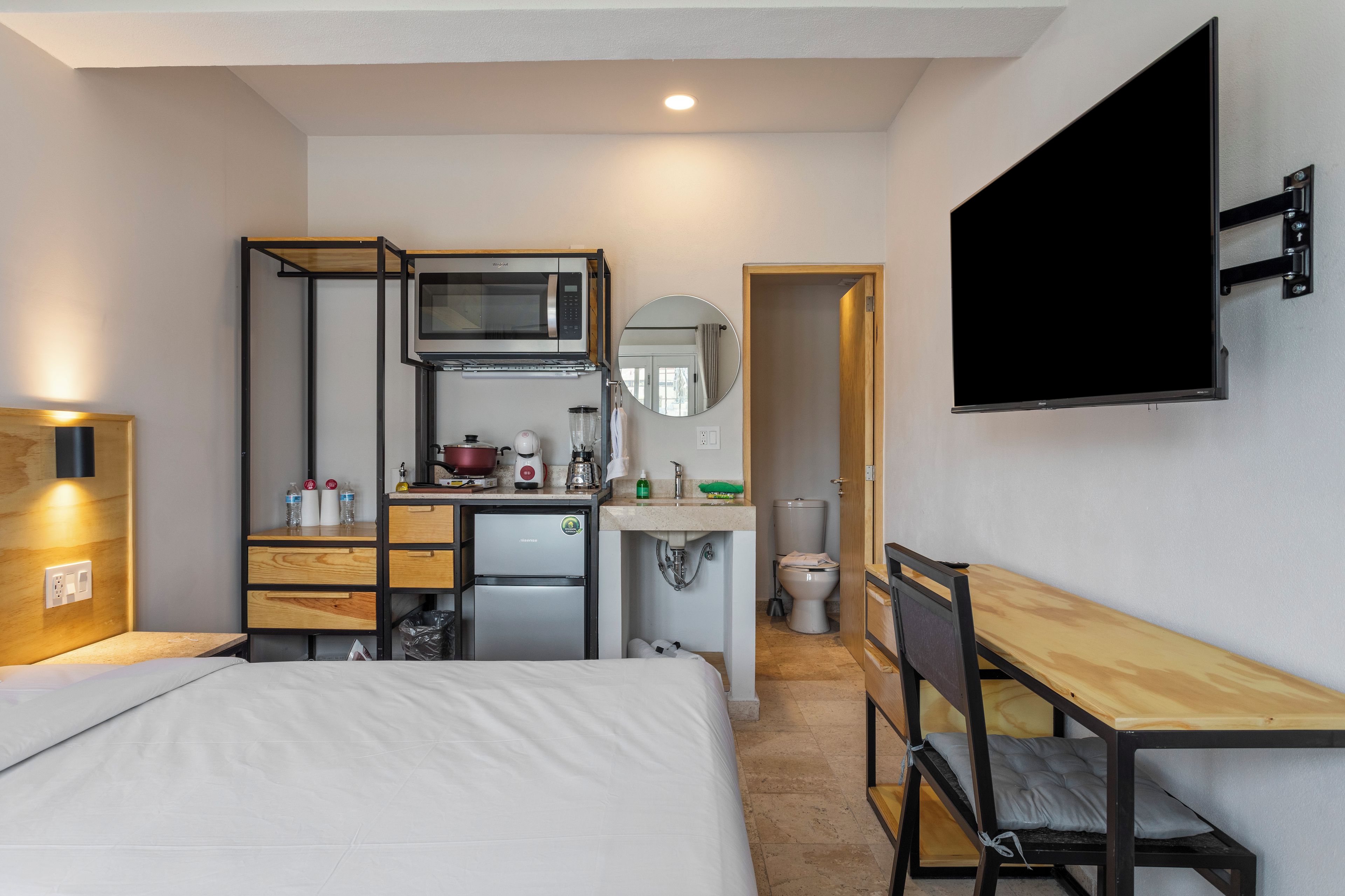 A compact hotel room with a bed, a kitchenette, a small fridge, a microwave, a sink, a toilet, and a TV mounted on the wall.