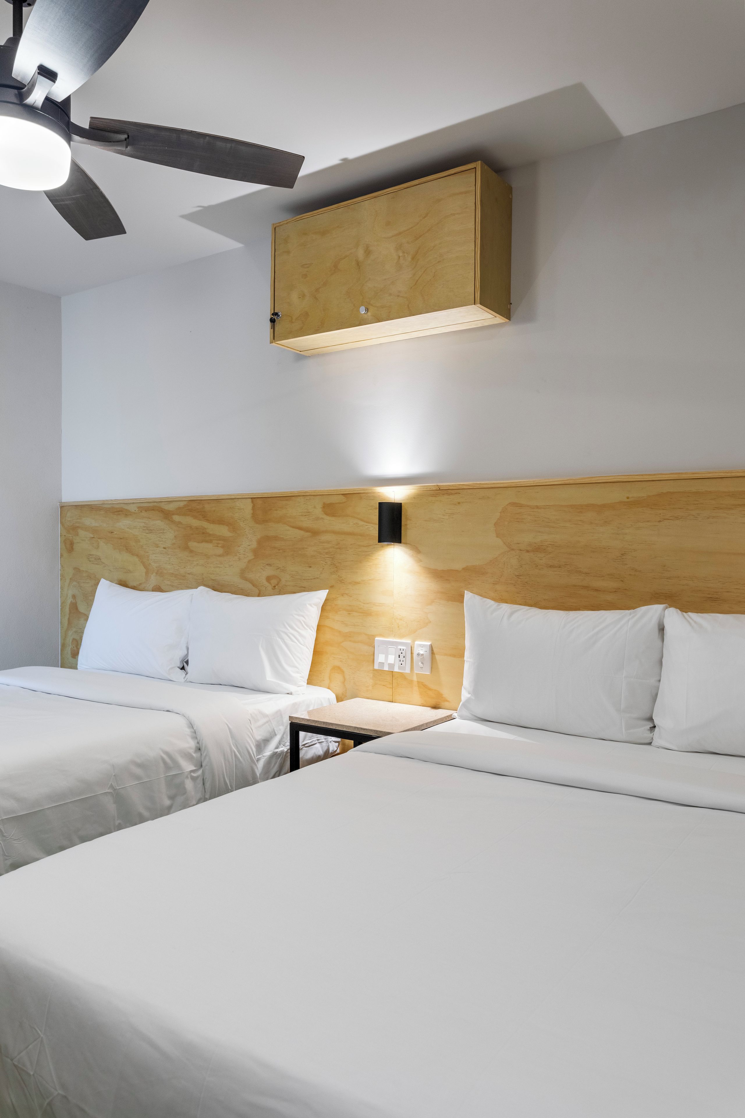Habitación de hotel con dos camas, cabecero de madera, ventilador de techo e iluminación montada en la pared.