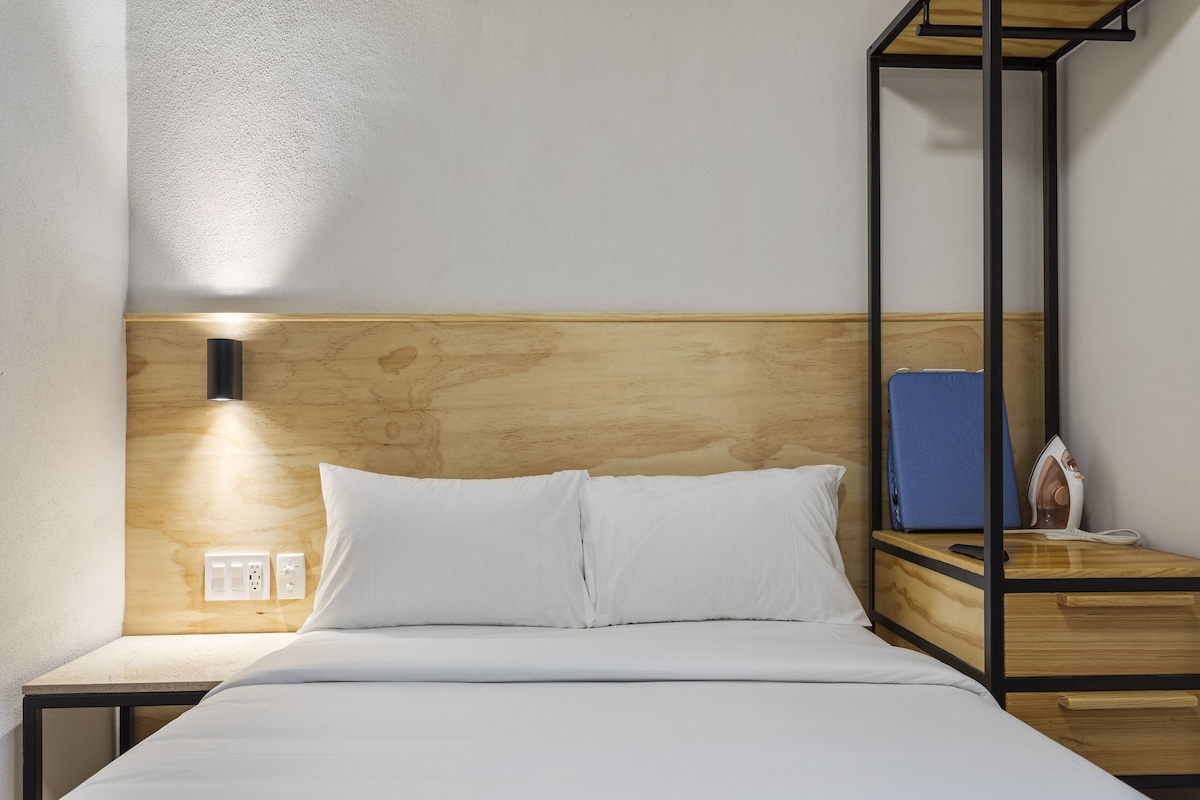 Habitación de hotel con cabecera de madera, cama con ropa blanca, mesita de noche con lámpara y un libro azul sobre la mesita.