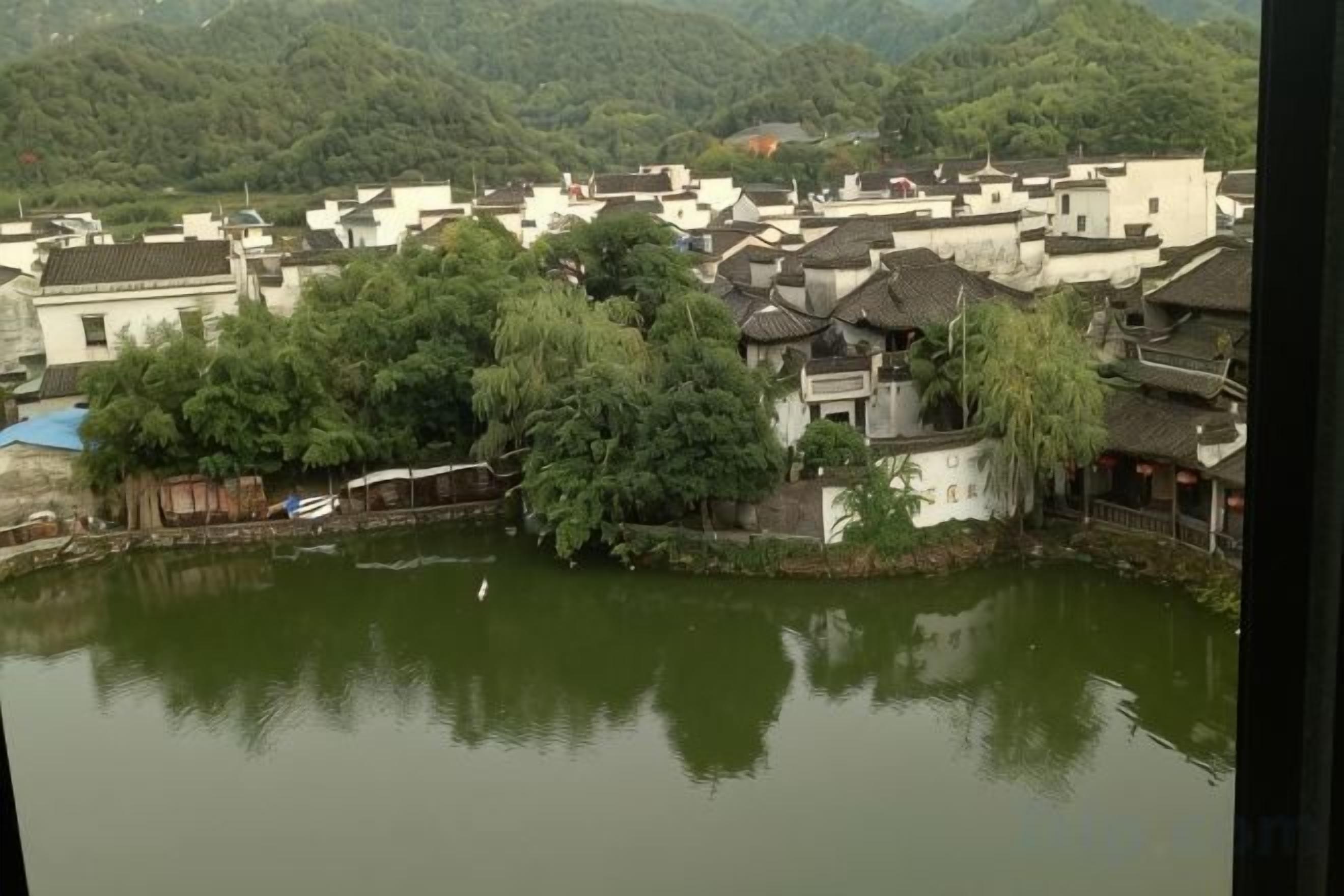 A traditional village with buildings clustered around a central water body, surrounded by greenery and hills.