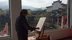 Une personne est en train de peindre devant une fenêtre, avec vue sur des bâtiments traditionnels et des montagnes.