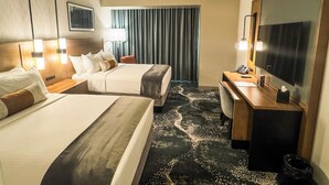 A hotel room with two beds, a desk, a TV, and a large window with curtains.