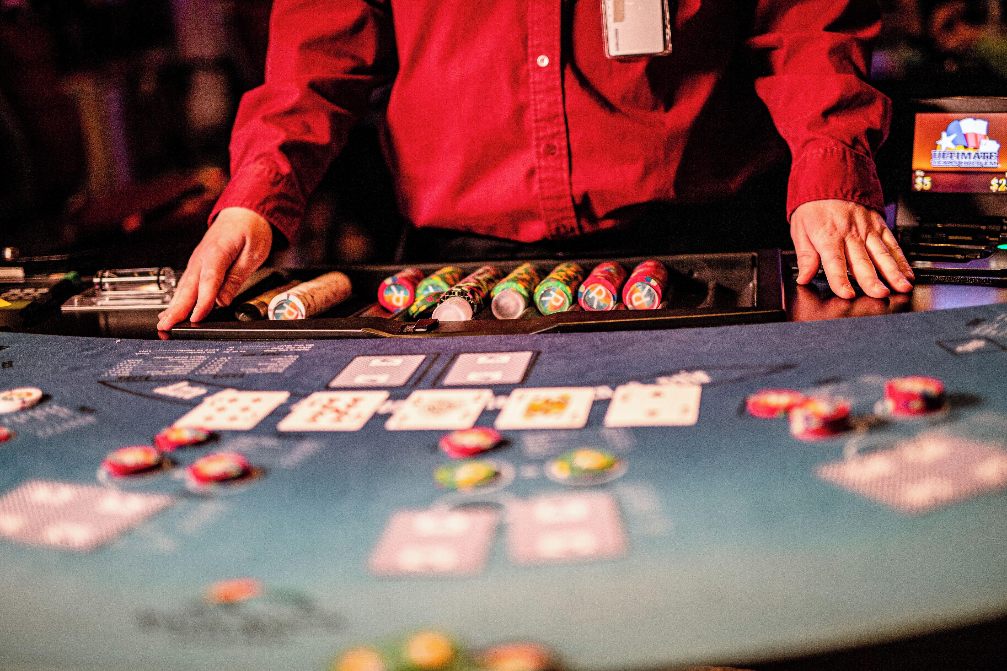 A blackjack table with cards and chips, a person in a red shirt, and a digital screen displaying game options.
