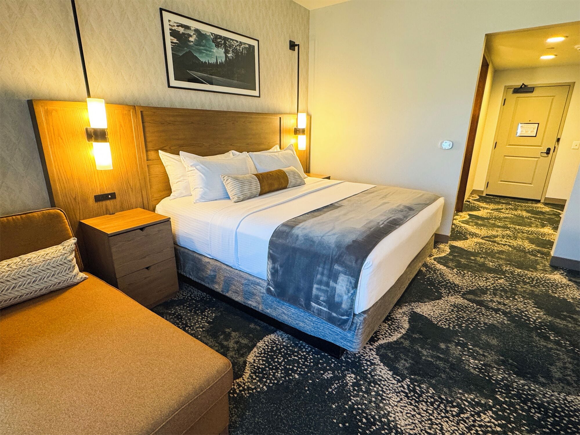A hotel room with a large bed, a nightstand, a chair, and a door leading to another room.