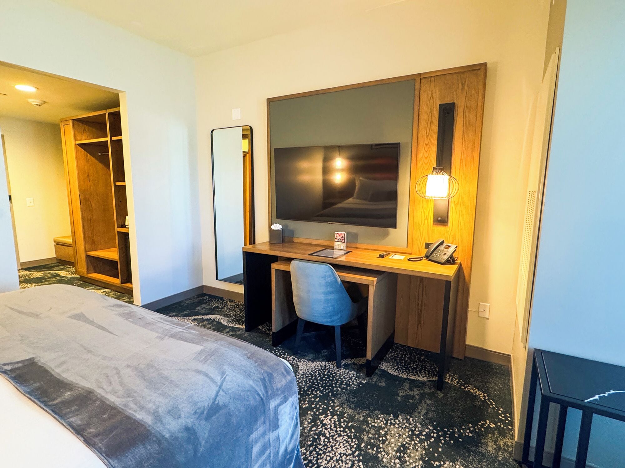 A hotel room with a bed, a desk with a chair, a large mirror, a flat-screen TV, a telephone, and a small table with a tissue box.