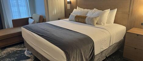 A hotel room with a large bed, a bedside table, a nightstand, a chair, and a window with curtains.