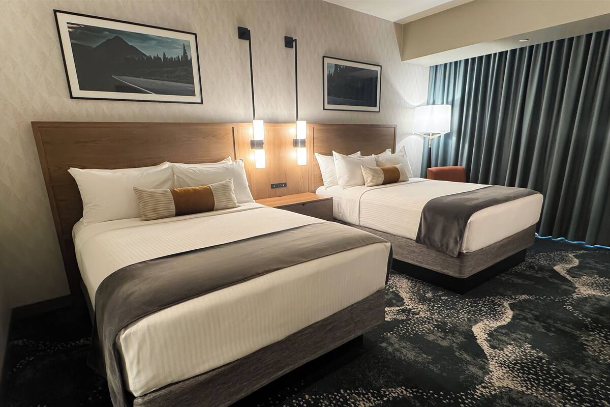 Ein Hotelzimmer mit zwei Betten, einem Kopfteil, Nachttischlampen, einem Nachttisch, einem Sessel, einem großen Fenster mit Vorhängen und gerahmten Bildern an der Wand.