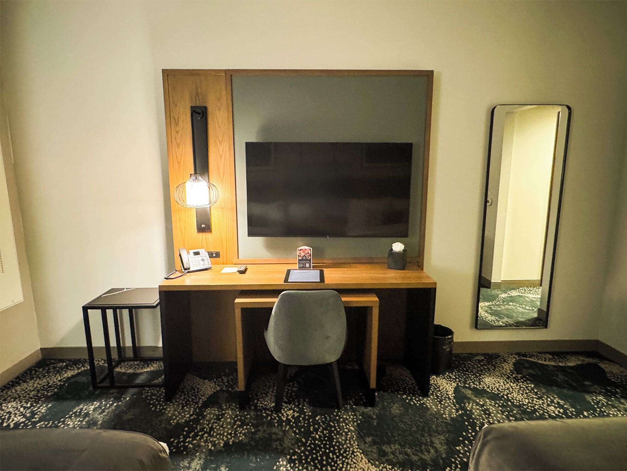 Habitación de hotel con un escritorio de madera, una silla, un teléfono, una mesita, un espejo y una alfombra estampada.