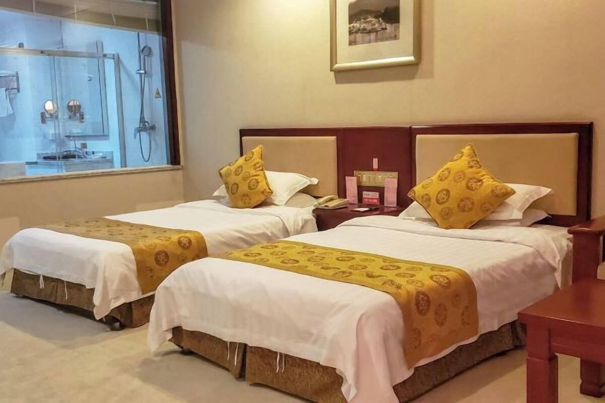 Habitación de hotel con dos camas, cada una con cubrecama blanco y manta amarilla estampada. Tiene cabecera de madera, mesita de noche con lámpara y un cuadro enmarcado en la pared.