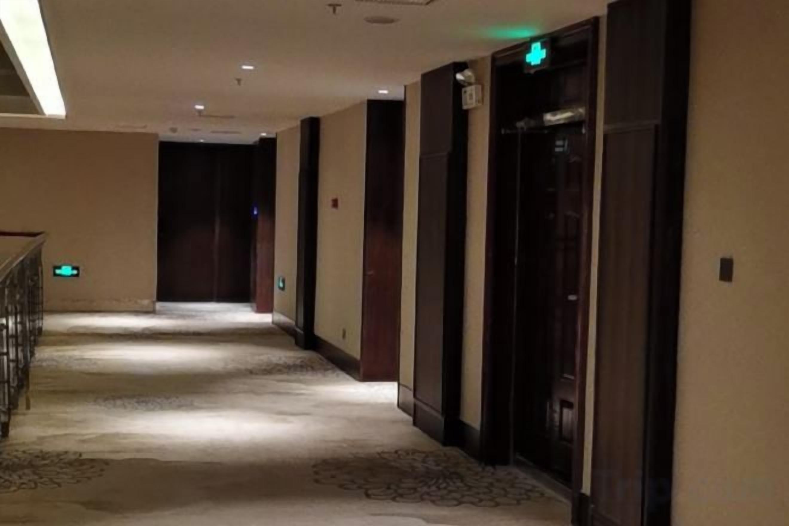 Un pasillo de hotel con piso alfombrado, puertas de madera y un letrero de salida verde.