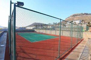 A tennis court enclosed by a green fence with a clear sky and a hilly landscape in the background.
