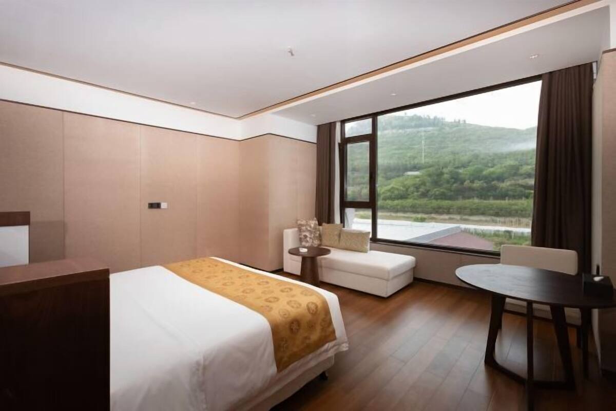 Una habitación de hotel moderna con una cama grande, un sofá pequeño, una mesa redonda y vistas a un paisaje verde a través de un ventanal amplio.