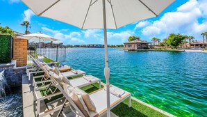 Une terrasse au bord de l’eau, dotée de chaises longues et de parasols, une piscine, et une vue sur la mer et les bâtiments en arrière-plan.