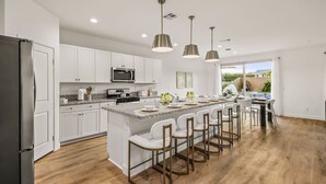 A modern kitchen with white cabinetry, a central island with bar stools, a stainless steel refrigerator, and pendant lighting. - 15 Bed, Glow Gameroom, Putting green, Pool & BBQ (Coachella)