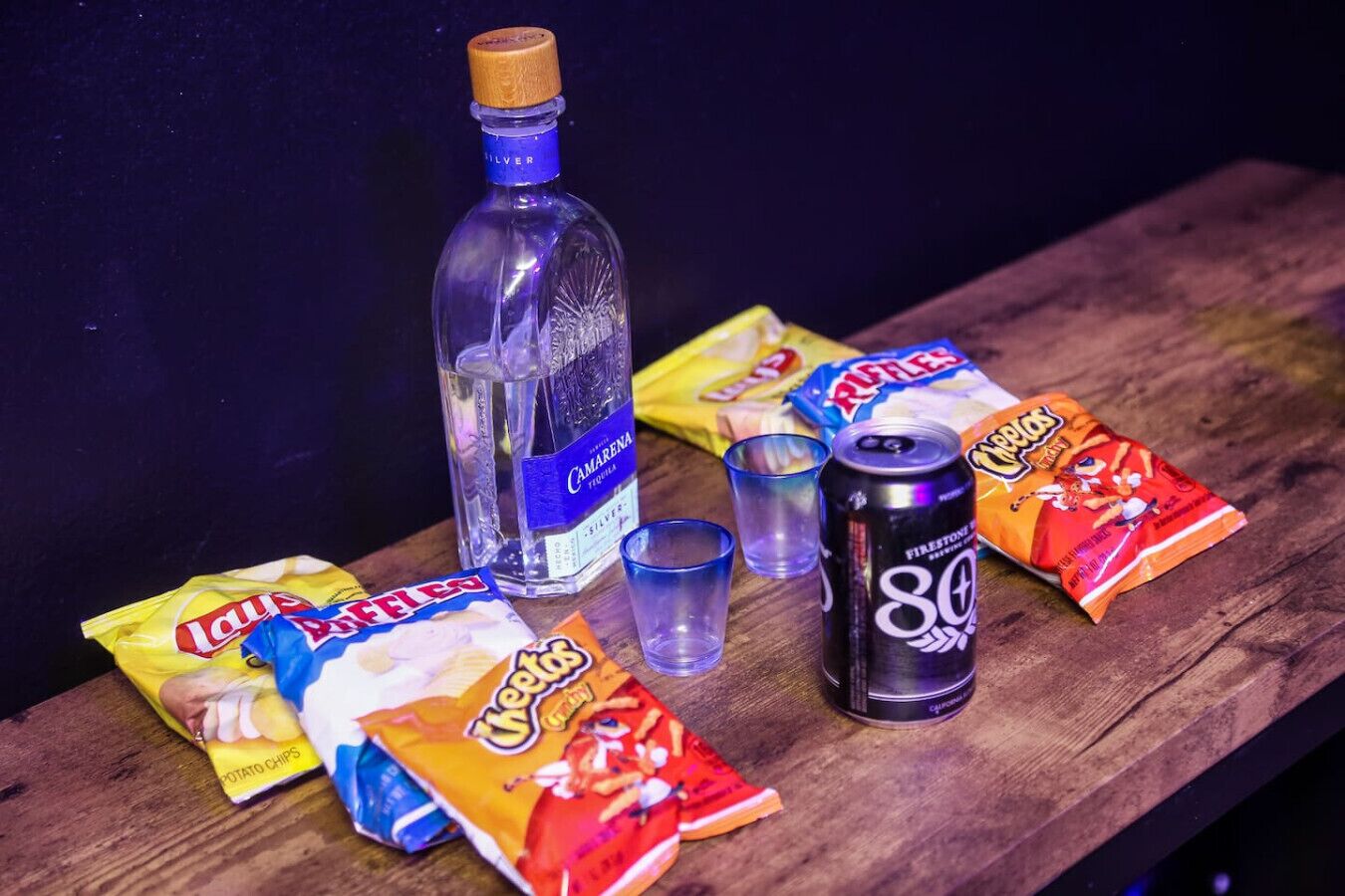 Een houten tafel met een fles Camarena tequila, twee glaasjes, een blikje 80s en zakken Lay's chips.