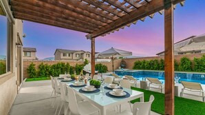 An outdoor patio with a dining table set for six, surrounded by white chairs, under a wooden pergola. A swimming pool is visible in the background. - 15 Bed, Glow Gameroom, Putting green, Pool & BBQ (Coachella)