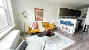 En moderne stue med en gul sofa, en grå lænestol og et sofabord. Der er et køkkenområde med en bar og blå stole.