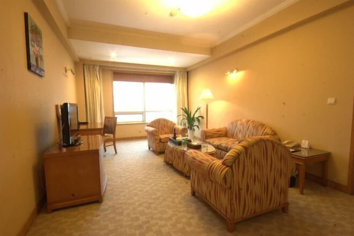 Quarto de hotel com sofá, poltrona, televisão, abajur e janela com cortinas.