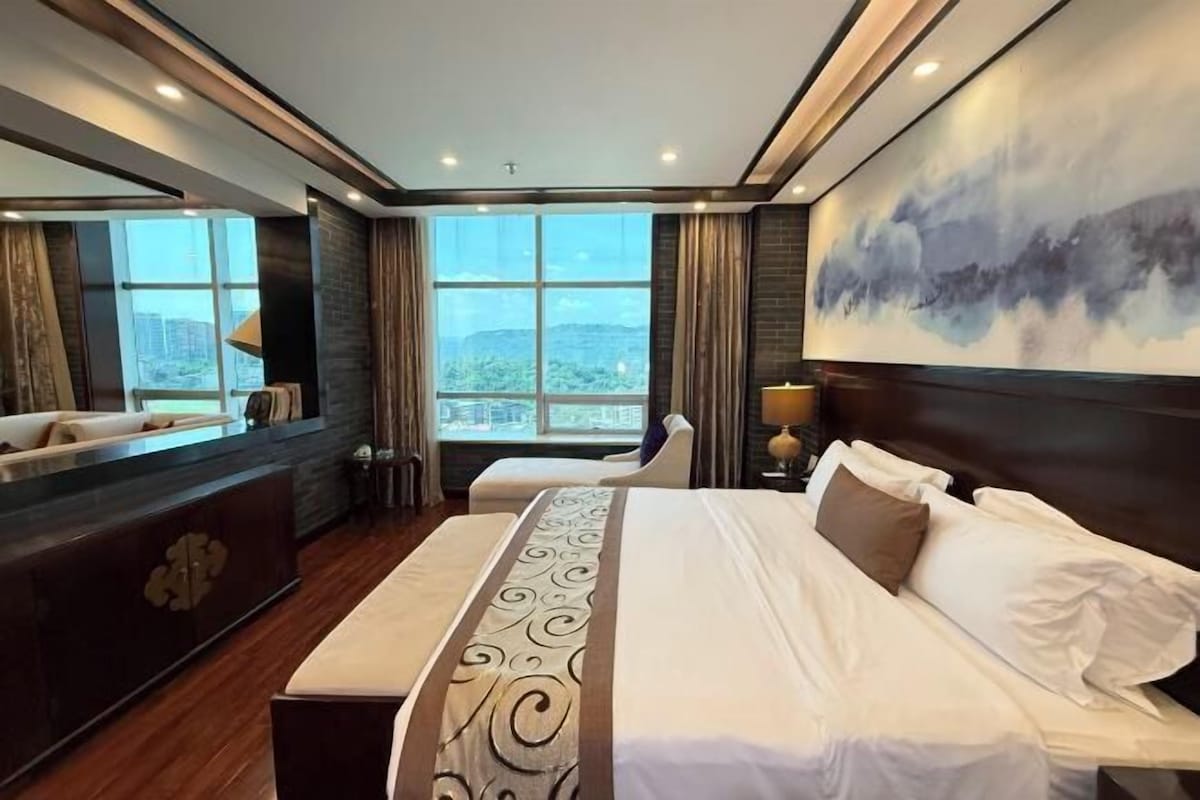 Quarto de hotel com cama grande, área de estar, escrivaninha e uma ampla janela com vista para a cidade.