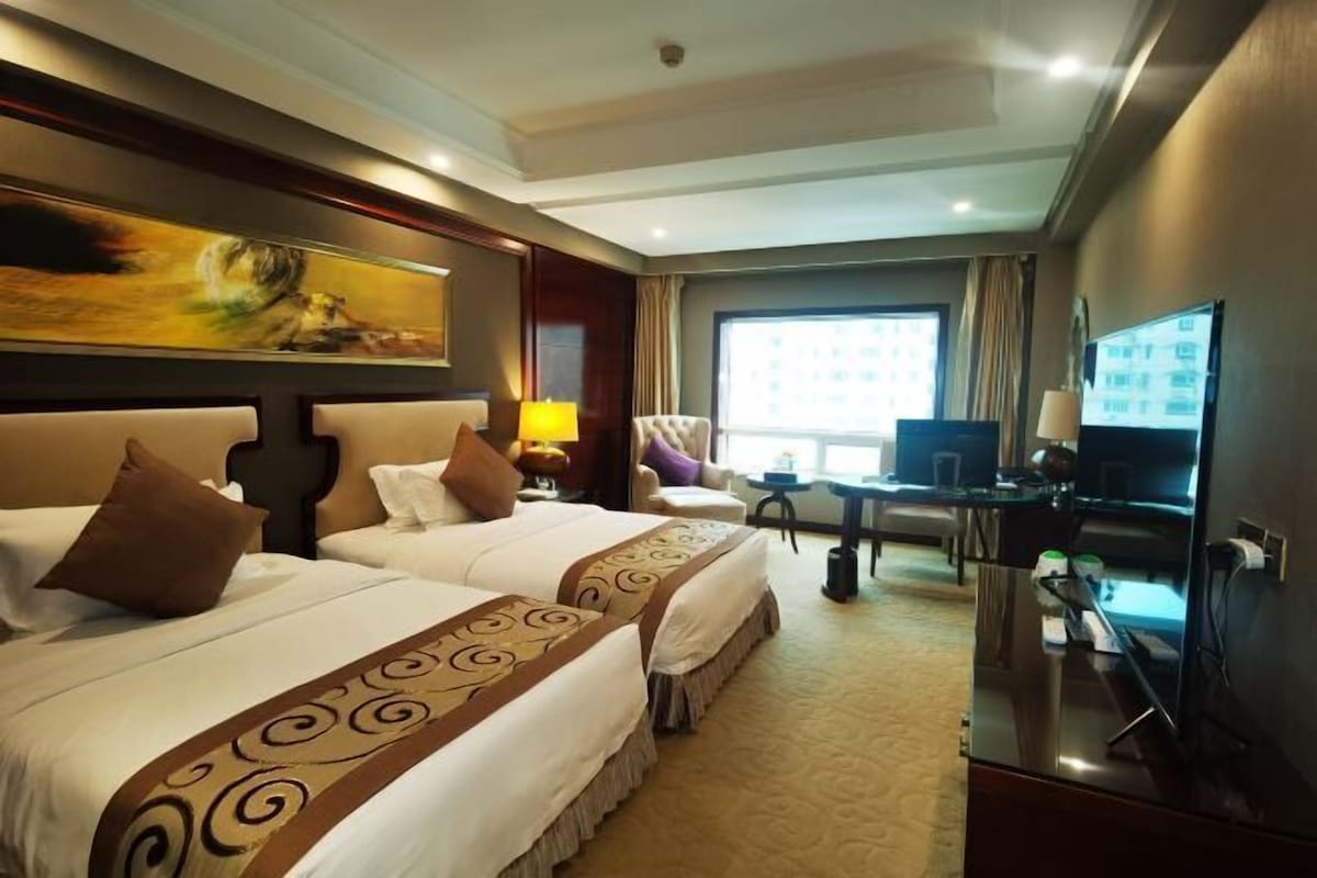 Quarto de hotel com duas camas, uma escrivaninha, uma televisão e uma janela grande.