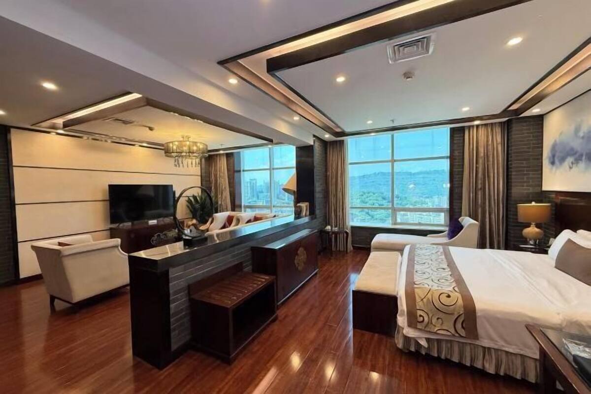 A modern hotel room with a large bed, a seating area with a TV, and a view of the city.