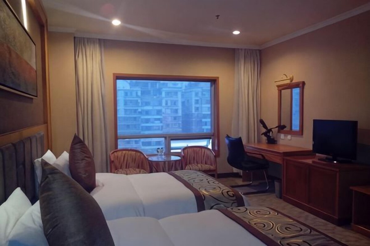 A hotel room with two beds, a desk, a chair, a television, and a window with a view of buildings.