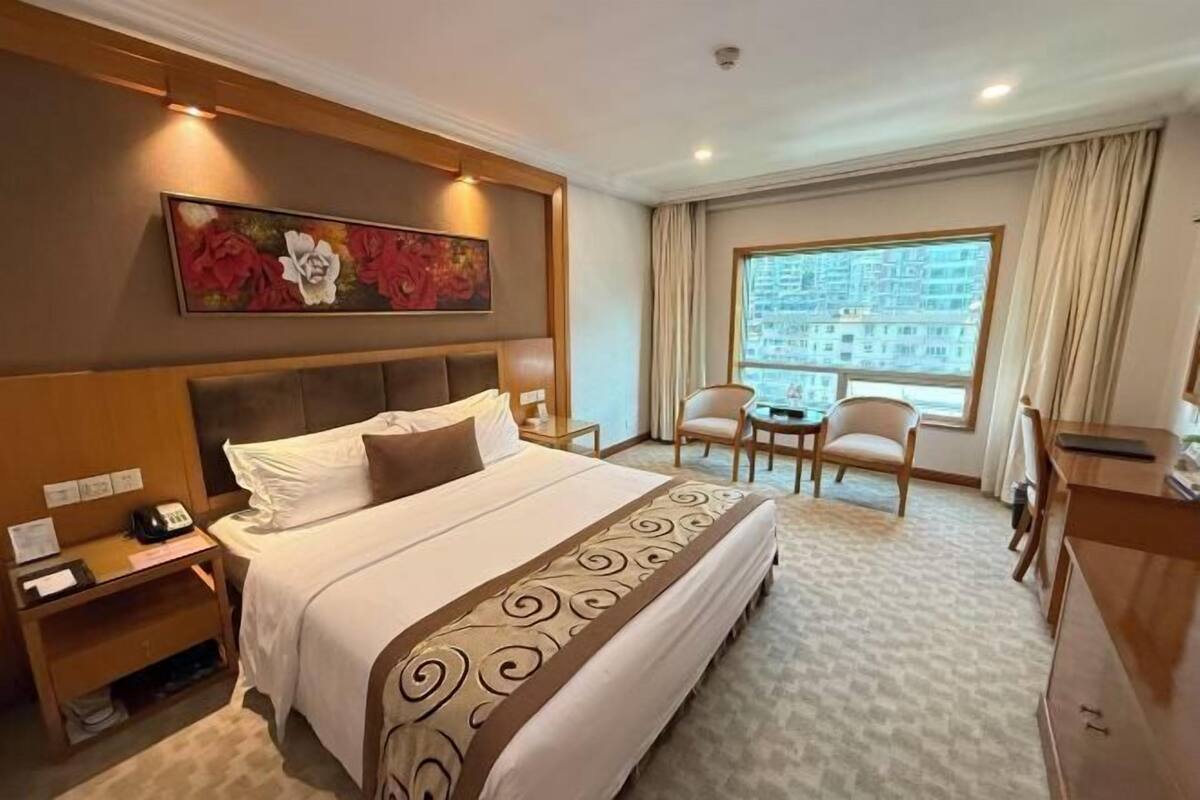 A hotel room with a large bed, a desk, a chair, a small table, and a window with a city view.