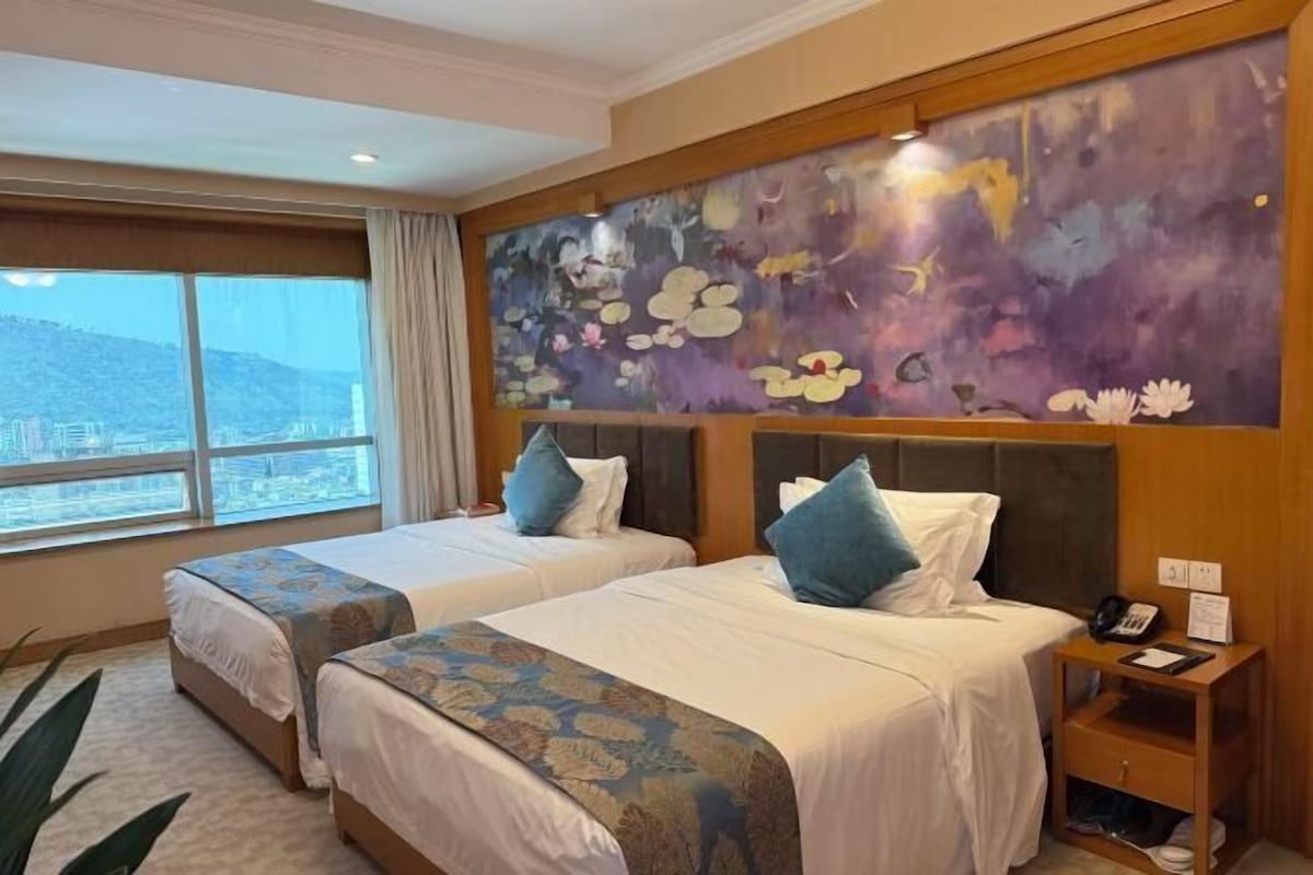 A hotel room with two beds, a large window with a city view, a floral mural on the wall, and a bedside table with a phone and a notepad.