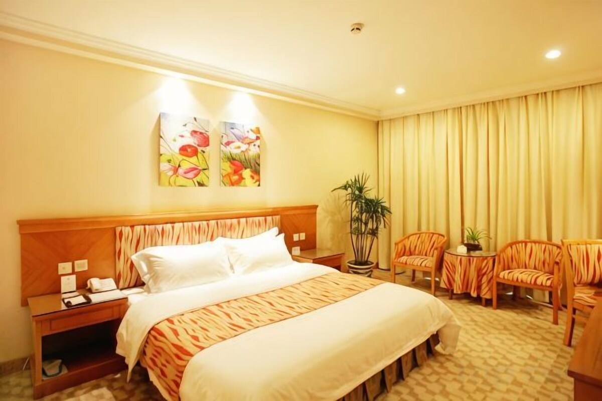 A hotel room with a large bed, two bedside tables, a nightstand, a potted plant, and a seating area with two chairs and a small table.