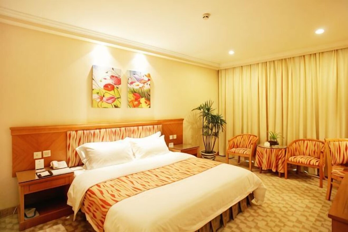 A hotel room with a large bed, two bedside tables, a nightstand, a potted plant, and a seating area with chairs.