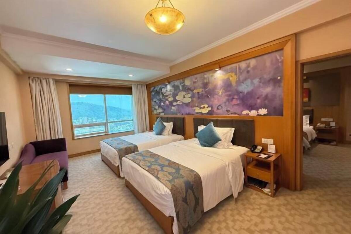 A hotel room with two beds, a large window, a painting on the wall, and a plant.