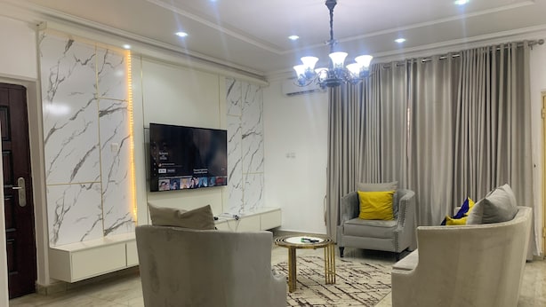 A modern living room with a flat-screen TV mounted on a marble wall, two armchairs, a small round table, and a grey sofa with a yellow pillow. - 4mins to Aso-Rock Villa, King Bed, Chef, Free Cleaning, 24/7 Electricity  (Abuja)