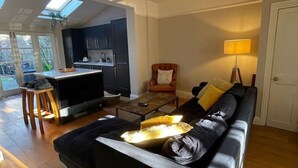 A modern living room with a black sofa, a coffee table, and a TV. There is a kitchen area with dark cabinets and a dining table. A lamp is on the right side, and a door is visible on the right. - A sun-kissed, cozy family home. (London)
