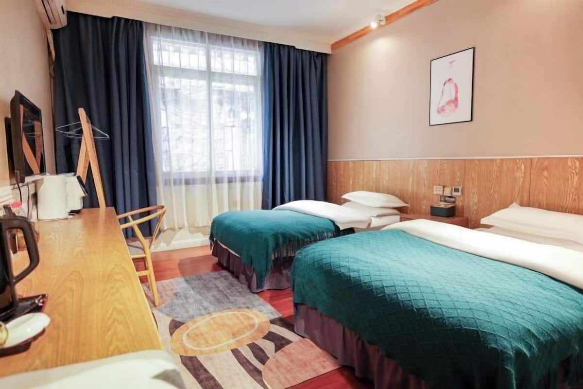 A hotel room with two beds, a desk with a chair, a radiator, and a window with curtains.