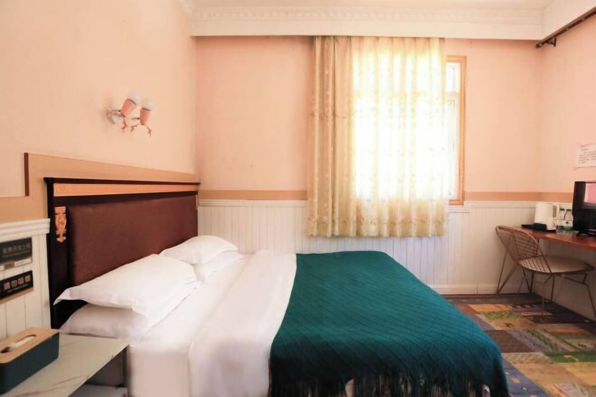 A hotel room with a bed, a desk, a chair, a window with curtains, and a wall-mounted telephone.