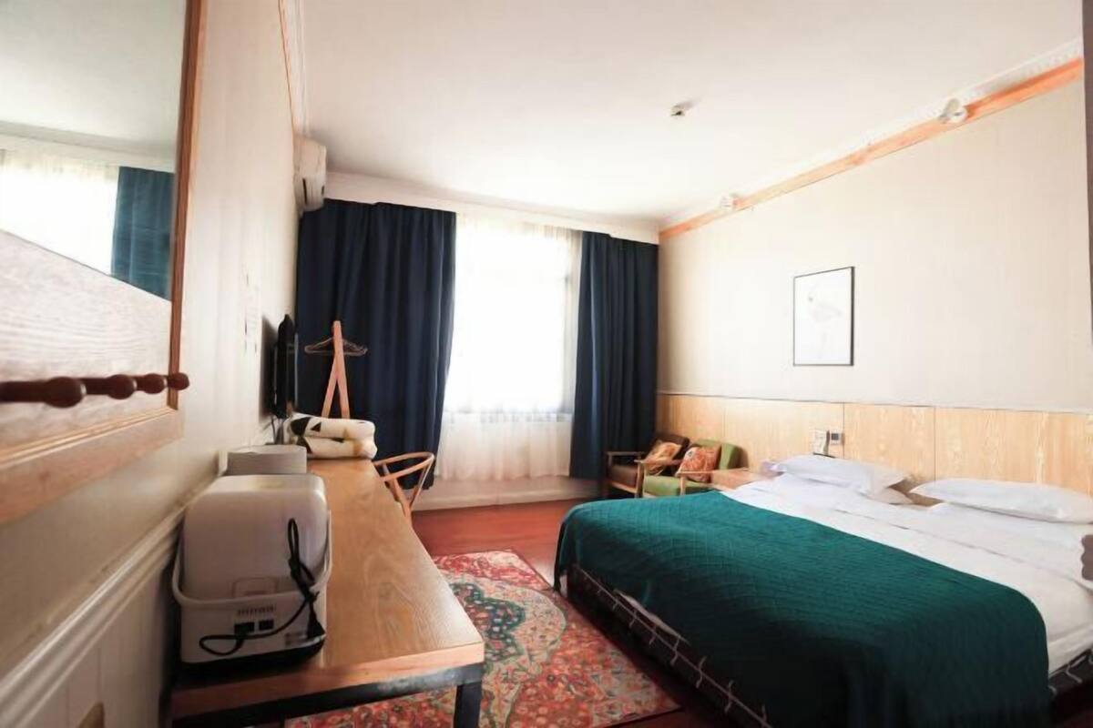 A hotel room with a bed, a desk with a coffee maker, a chair, a window with curtains, and a wall-mounted phone.